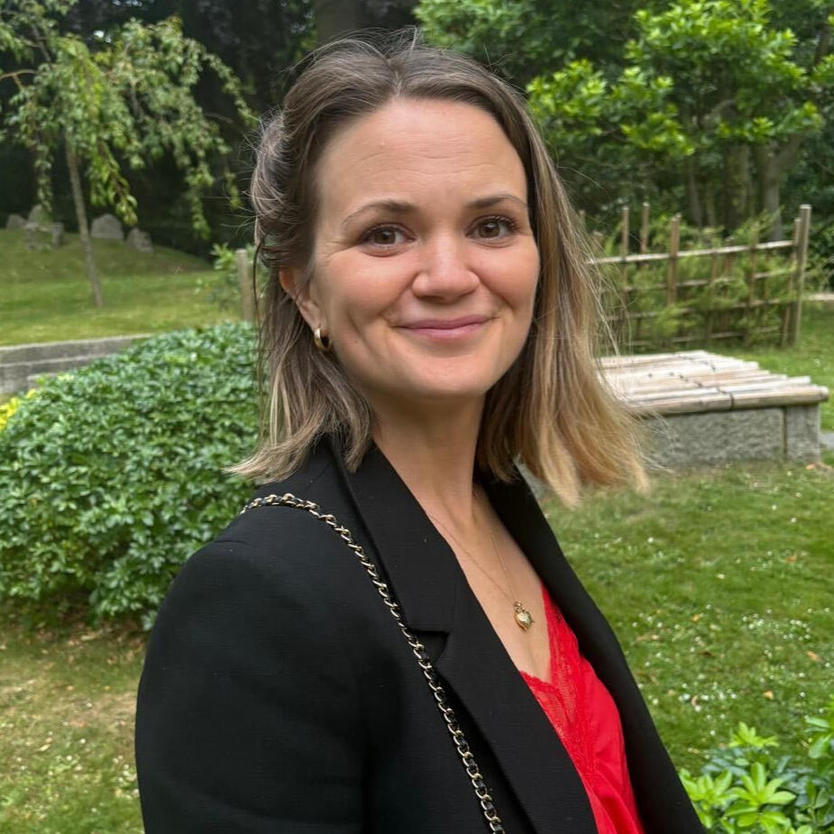 Kimberley, a white woman with long blonde hair, is smiling and standing in front of a large wreath decorated with fairy lights. She wears a warm cream-coloured coat.
