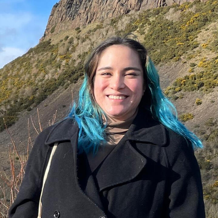 Rho, a mixed East Asian femme, smiles in front of the rocky slopes of the Salisbury crags. They have long bright blue hair and wear a black peacoat.