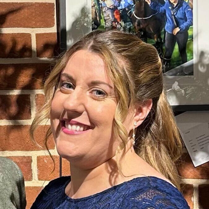 Maria, a white woman with blonde hair mostly tied back and two ringlets framing her face, smiles at the camera. She is wearing a lacy blue top and standing a sunny spot.