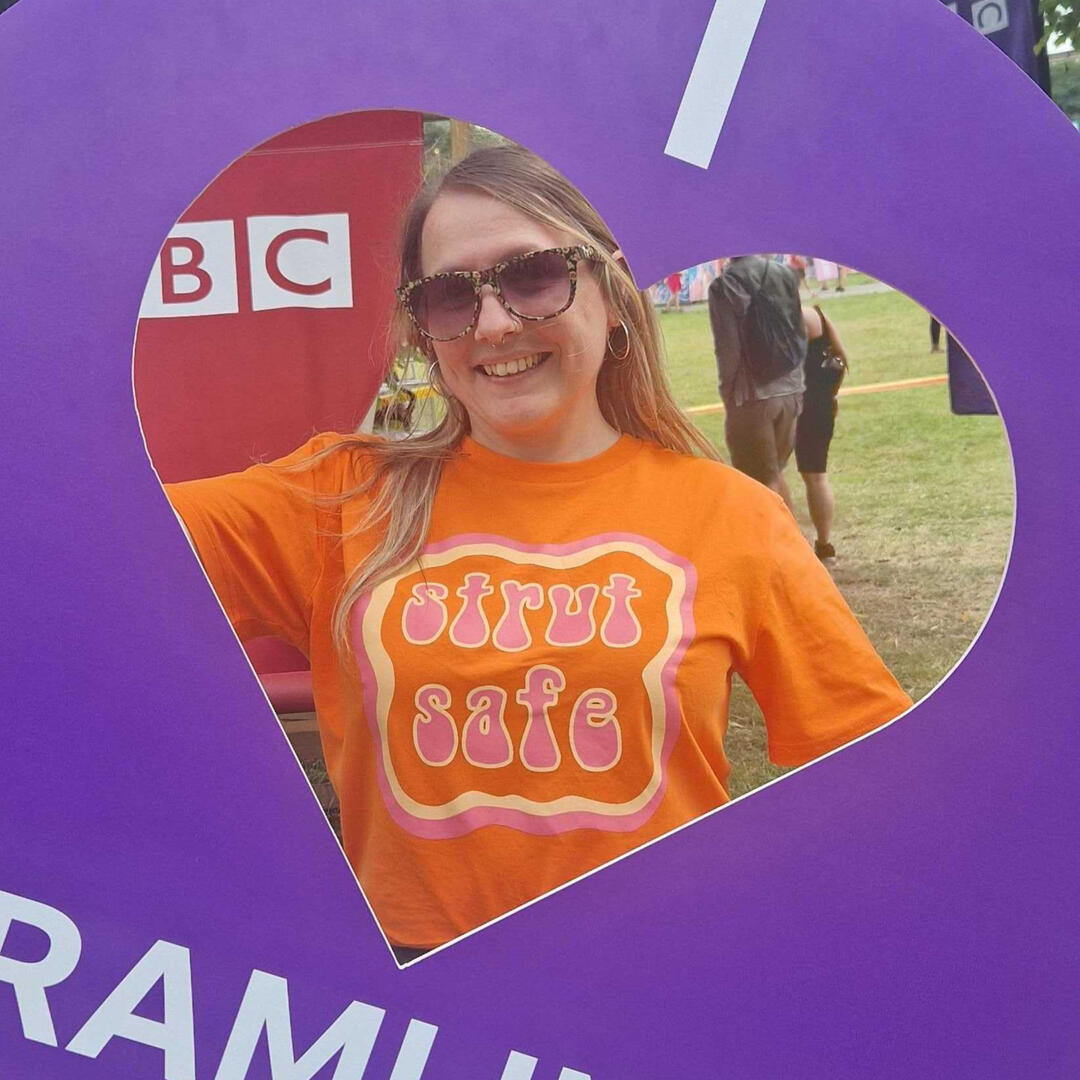 Nia, a white woman with blonde hair is posing inside of a purle heart-shaped frame. She is wearing a bright orange Strut Safe t-shirt and sunglasses.
