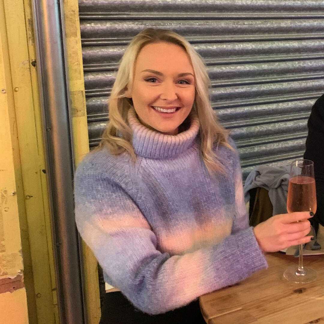 Laur, a white woman wearing a thick pink and purple jumper, is sitting at a small table. She has blonde curly hair and is holding a sparkling pink drink. She smiles widely for the camera.